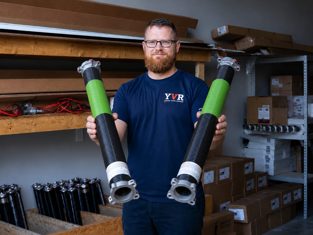 A YVR Garage Door Springs technician in the shop holding two heavier-gauge 25,000-cycle high-cycle torsion springs, identifiable by their green protective wrappers.