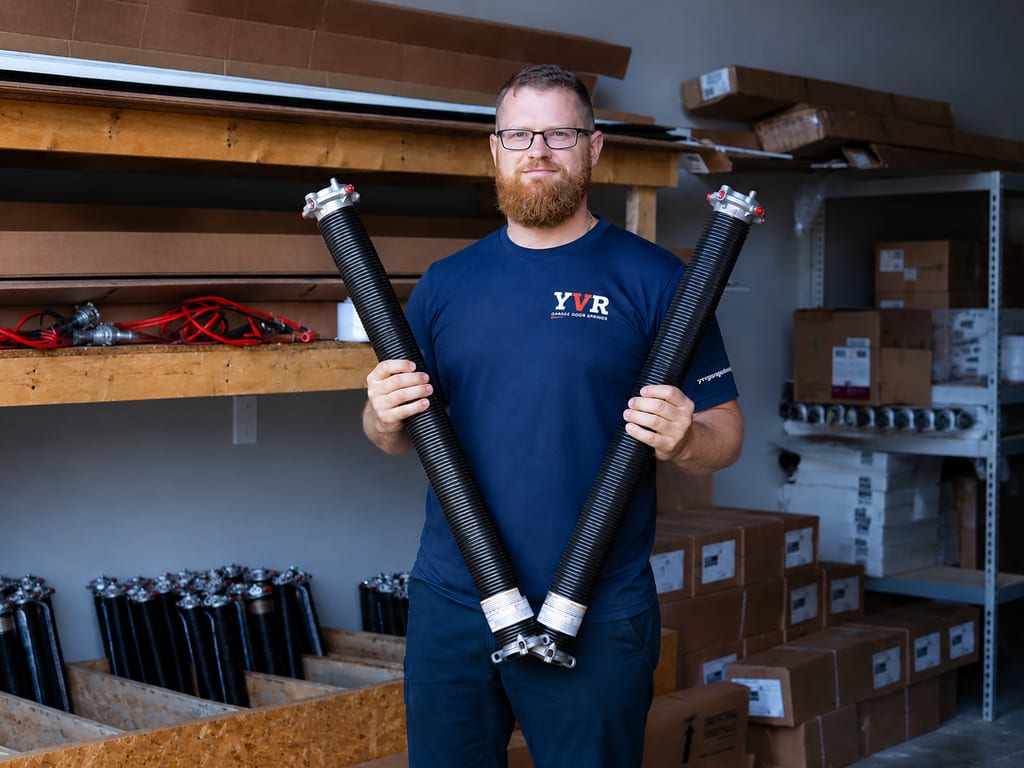 A YVR Garage Door Springs technician in the shop holding two matching black oil-tempered residential torsion springs.