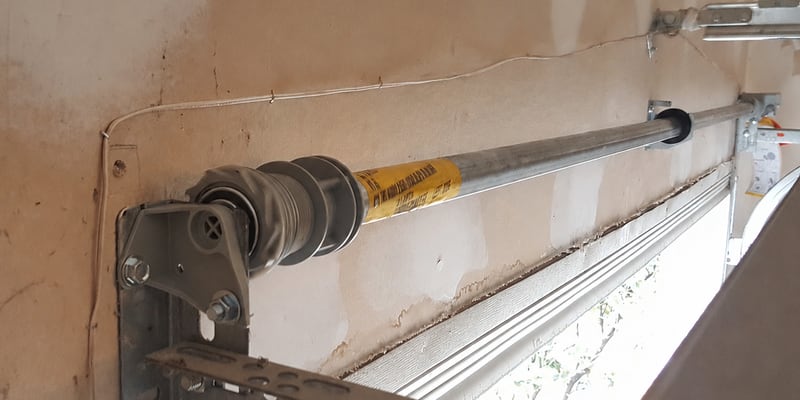 A Wayne Dalton TorqueMaster system installed on a residential garage door header — the proprietary sealed steel tube hides the torsion spring inside, with the original yellow factory warning label still attached to the winder cone.